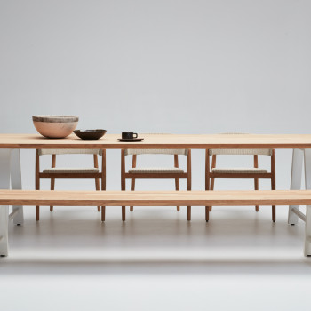 A modern dining setup featuring the Vincent Sheppard Matteo dining table with a solid wood top and white metal legs, paired with a matching Matteo bench. The arrangement is complemented by three woven dining chairs with wooden frames, and ceramic tableware adds a minimalist touch against a neutral backdrop.
