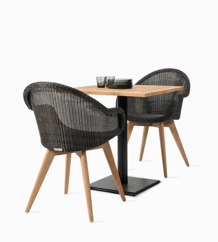The Vincent Sheppard Clark bistro table with a teak top and black metal base, paired with two Edgard dining chairs featuring a black woven seat and teak legs. A minimalist table setting with dark glassware adds elegance.