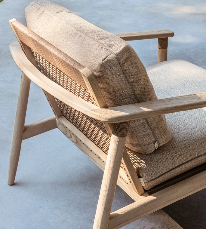 Close-up van de Vincent Sheppard David Lounge Chair, met een natuurlijk teakhouten frame, gevlochten rotan rugleuning en zachte beige kussens. De stoel staat op een betonnen buitenvloer in zacht zonlicht.