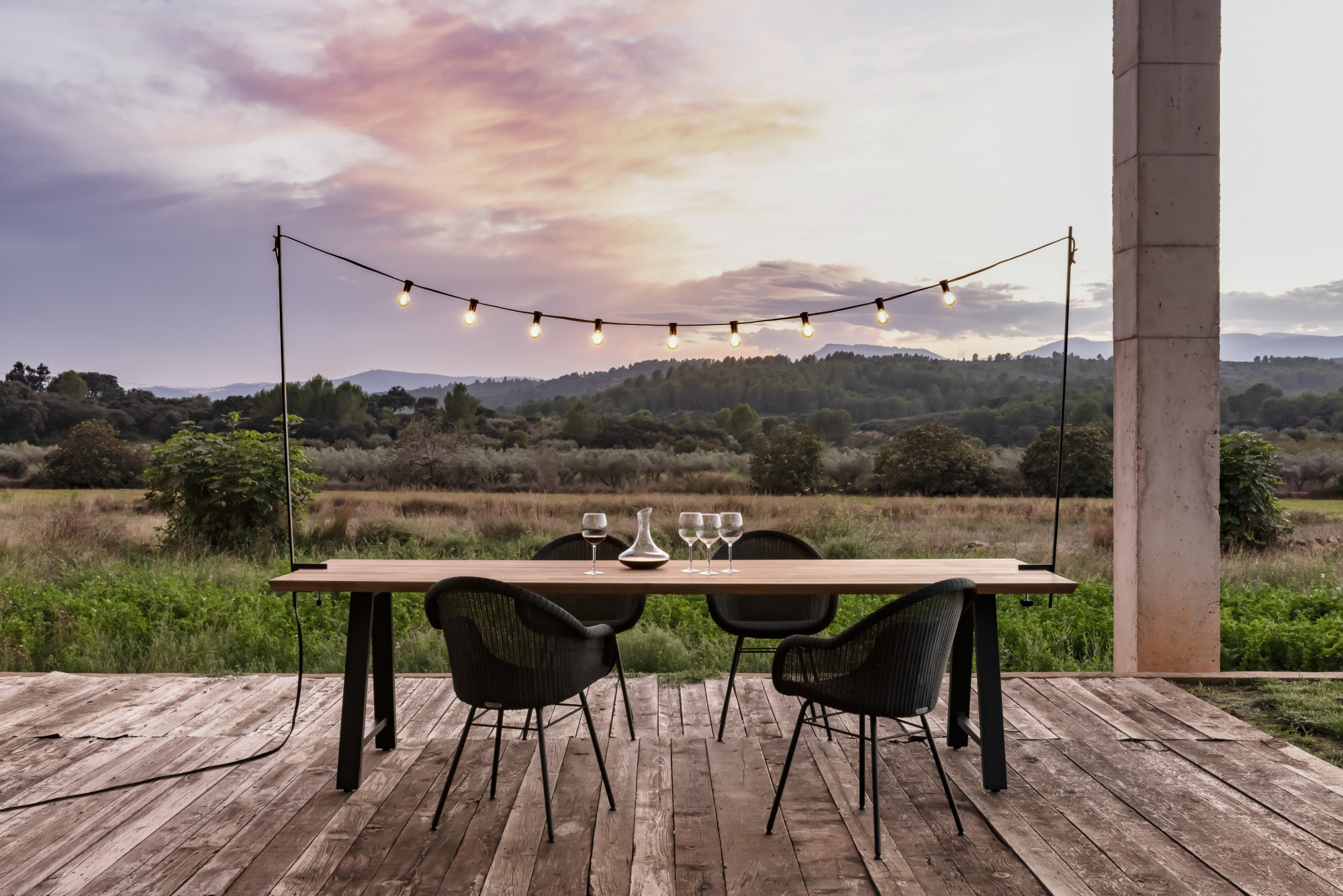 Coin repas extérieur avec les guirlandes lumineuses Light My Table de Vincent Sheppard, une table à manger Matteo en bois et des chaises Edgard noires. La scène surplombe un champ au coucher du soleil avec des nuages roses et violets.