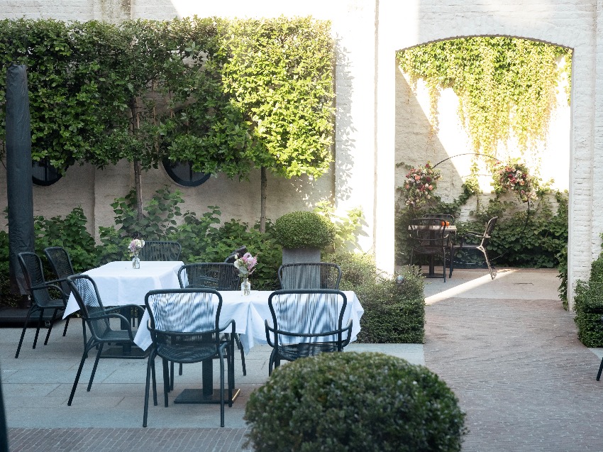 Espace de restauration extérieur à l'hôtel Cobergher à Courtrai avec des tables couvertes de nappes blanches, des chaises Loop et de la verdure environnante.