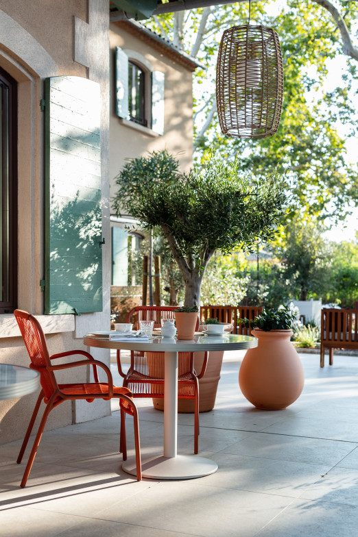 Espace extérieur à La Bastide Bourrelly avec une petite table ronde dressée pour deux, entourée de chaises de salle à manger Loop en terracotta, de plantes en pot et d'un grand olivier sous une lampe suspendue en osier.