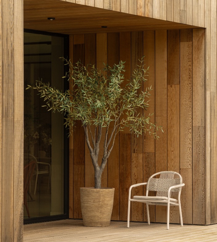 Outdoor area with a Vincent Sheppard Cleo lounge chair in dune white placed beside a large potted olive tree in a Vincent Sheppard Saya plant pot. The space features wooden cladding on the walls and decking
