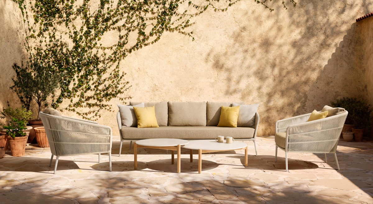 Vincent Sheppard Akari lounge set with a three-seater sofa and two armchairs featuring woven detailing and plush cushions, paired with Oda coffee tables with round tops and wooden legs, set in a Mediterranean-style outdoor patio.